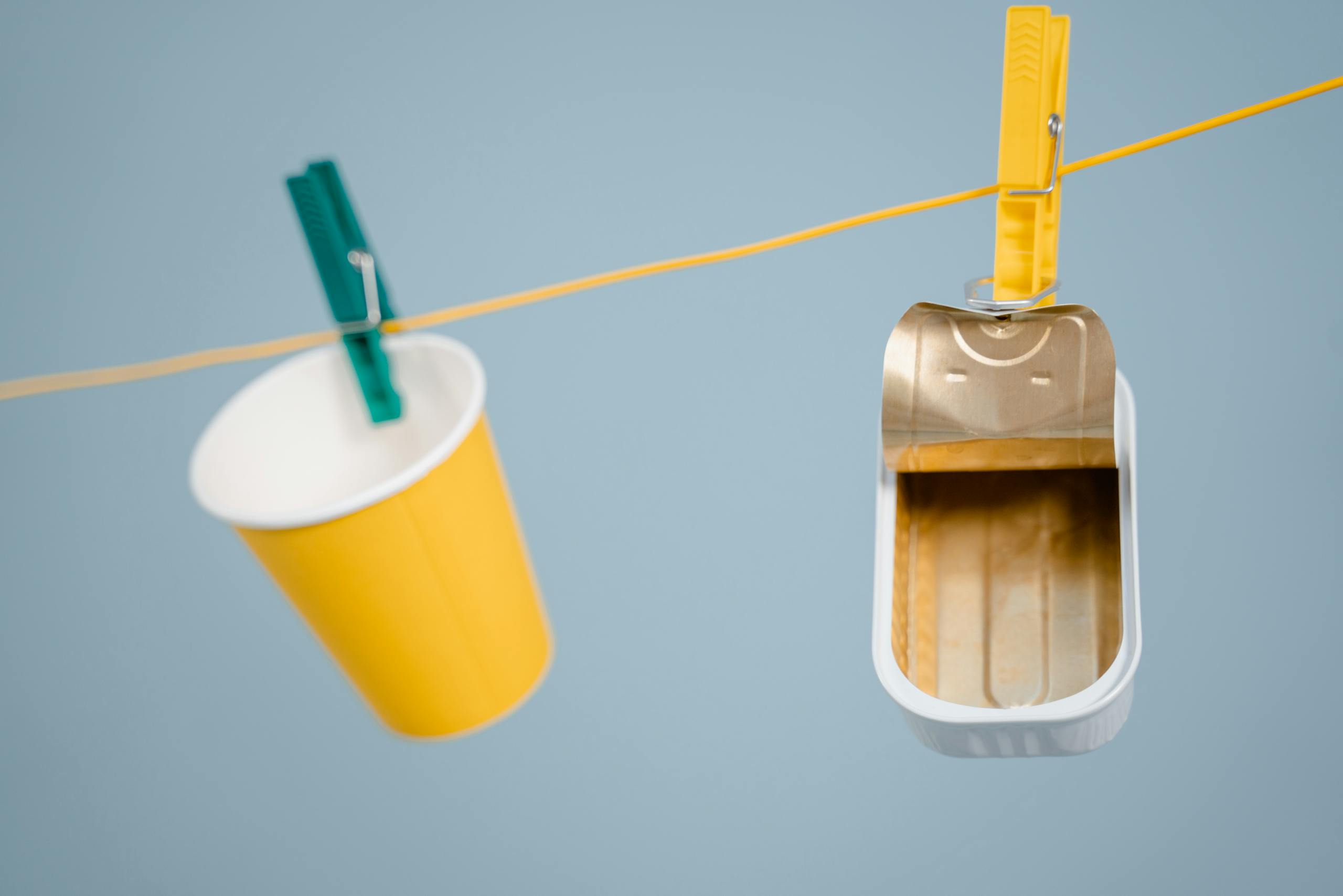 An artistic display of a yellow cup and can hanging on a clothesline with pegs against a blue background.